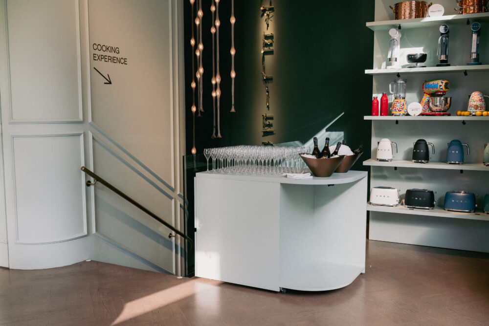 Interior of a showroom with white shelves displaying kettles, mixers, and barware; a sign reading 'COOKING EXPERIENCE' with an arrow to the left.