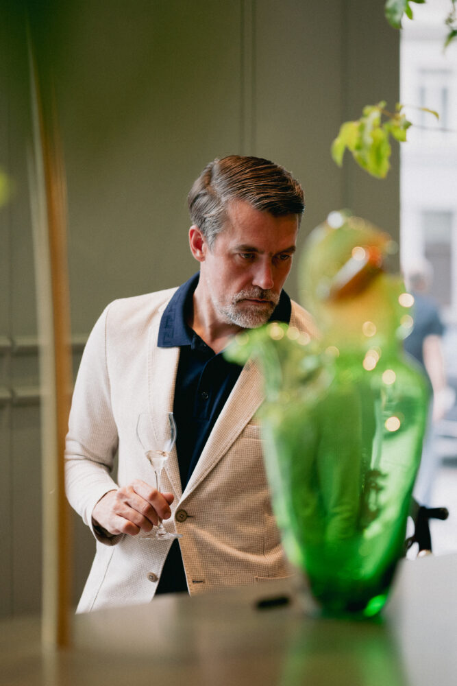 Man in a beige blazer holds a wine glass and looks downward, with a blurred green sculpture in the foreground indoors.