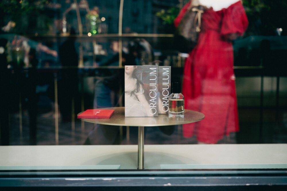 Display table in a shop window with two upright fashion magazines, a red booklet, and a perfume bottle; blurred red dress reflection in the glass.