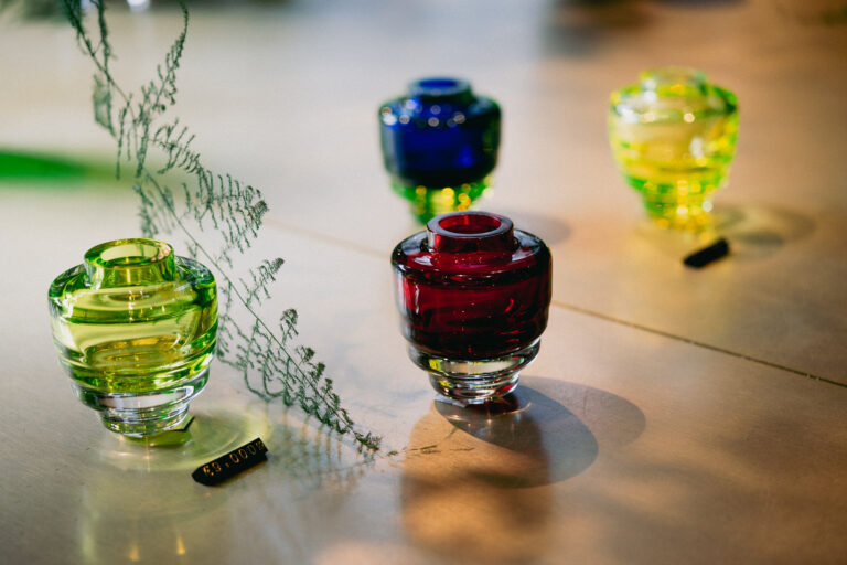 Colorful glass vases in green, blue, and red on a reflective surface with fern sprigs nearby.