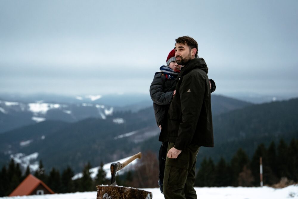 Man with beard holding a child in a dark jacket on a snowy mountain overlook with pine trees and hills in the distance