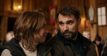 A man and woman sit close together, smiling softly at each other in a warm, rustic indoor setting.