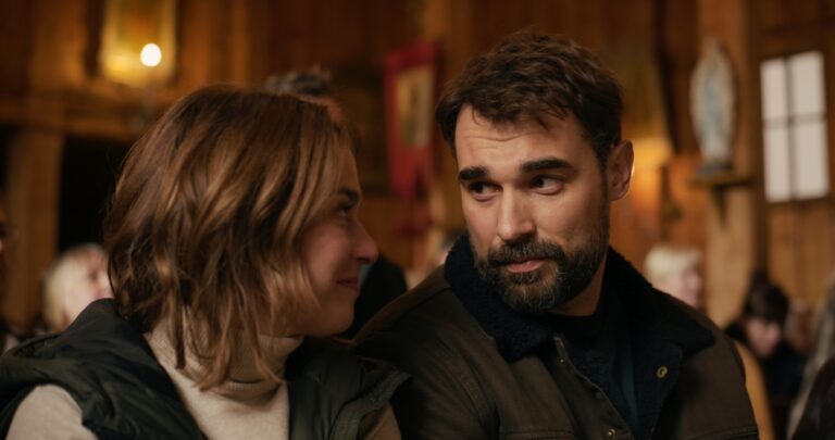 A man and woman sit close together, smiling softly at each other in a warm, rustic indoor setting.