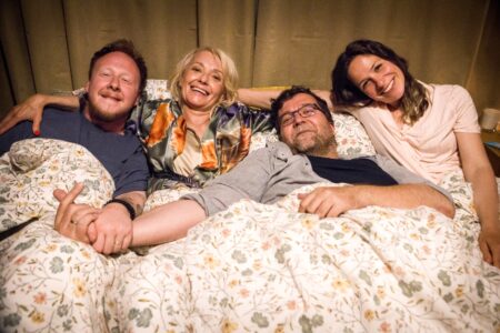Four adults lying in bed under a floral quilt, smiling at the camera.