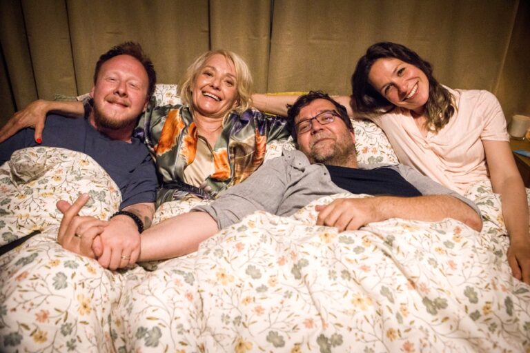 Four adults lying in bed under a floral quilt, smiling at the camera.