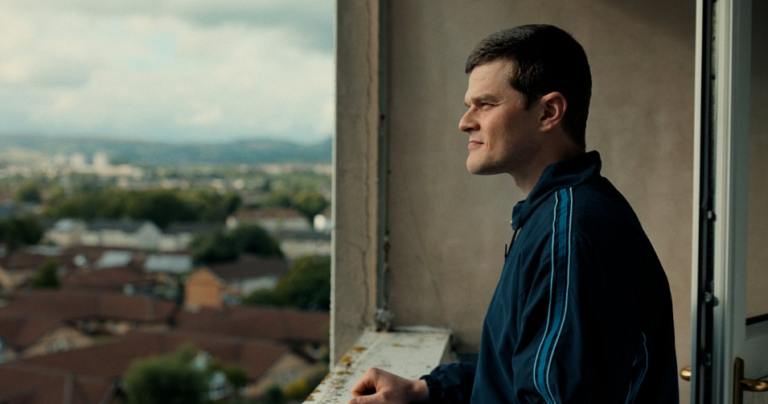 Man in a blue tracksuit on a balcony, gazing over a suburban cityscape to the left.