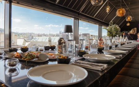 Long rooftop dining table set with plates, glasses and small dishes, overlooking a city skyline during the day.