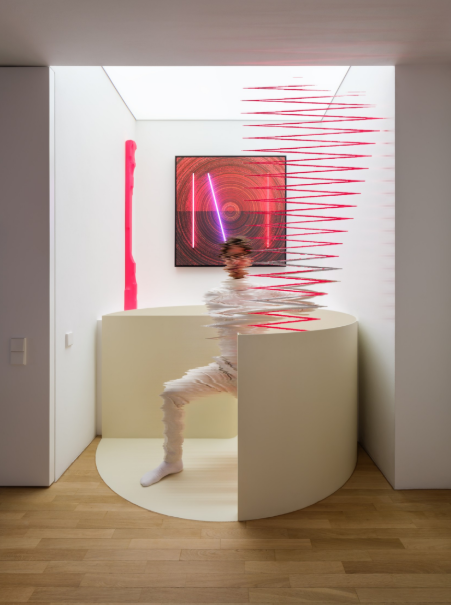 Person moving through a white circular partition in a gallery booth, creating motion blur, with neon pink light bars and a framed red artwork on the back wall.