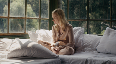 Woman sitting cross-legged on a bed by large windows, gazing downward with a thoughtful expression.