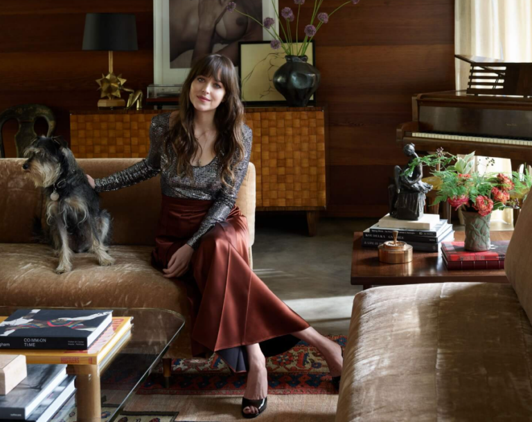 Woman with long brown hair sits on a tan sofa beside a small grey dog in a cozy mid-century living room with wood paneling, a piano, and decorative objects.