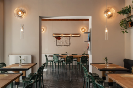 Modern cafe interior with wooden tables, green metal chairs, warm pendant lights, and potted plants; doorway leads to a larger back dining area.