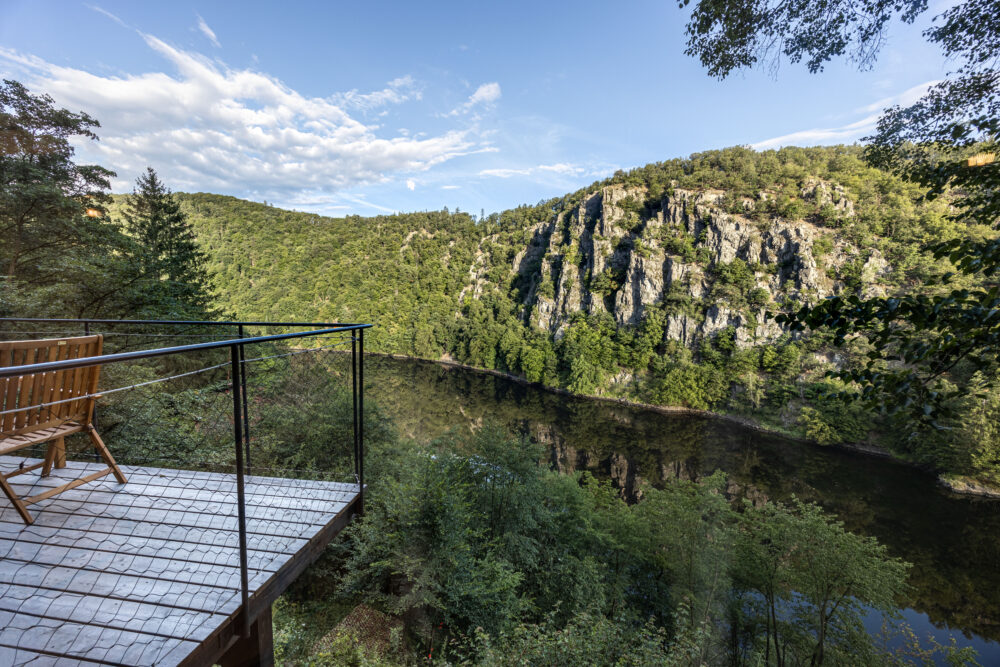 View from a wooden deck overlooking a forested canyon with rocky cliffs and a calm river below under a blue sky.