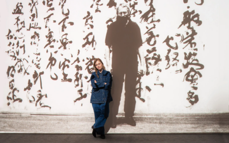 Person in denim outfit stands with arms crossed against a white wall covered in large black calligraphy; tall shadow looms behind them.