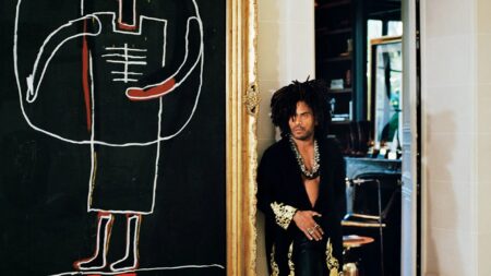 Man with an Afro hairstyle and open black shirt stands beside a gold-framed mirror, next to a large chalk drawing on a black wall.
