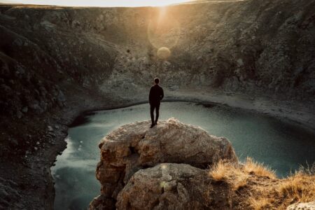 Person standing on a jagged rock overlooking a blue crater lake at sunset.