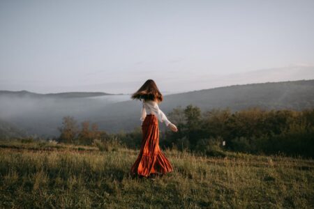 Woman in a white blouse and red maxi skirt walking through a misty hilltop meadow at sunset.