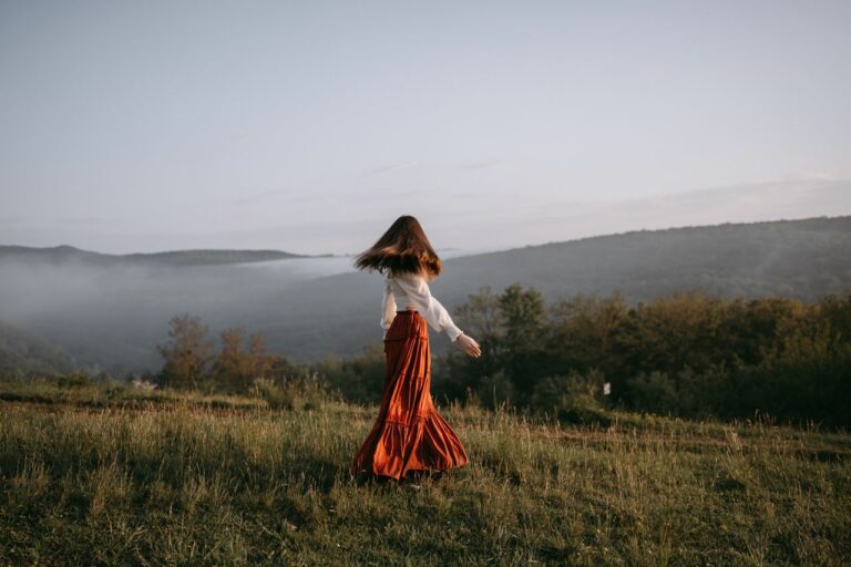 Woman in a white blouse and red maxi skirt walking through a misty hilltop meadow at sunset.