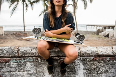 Person sitting on a weathered concrete wall at the beach with a skateboard across their lap, palm trees in the background.