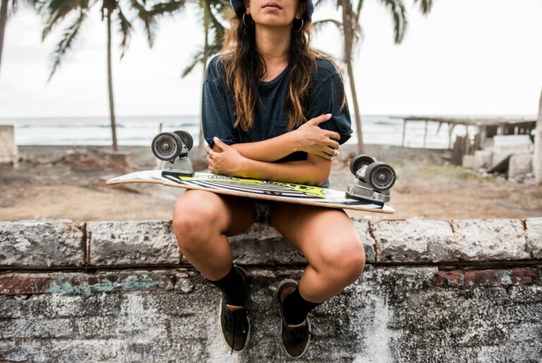 Person sitting on a weathered concrete wall at the beach with a skateboard across their lap, palm trees in the background.