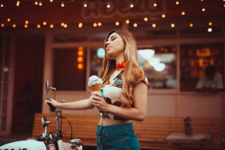 Young woman with long hair holding an ice cream cone beside a vintage motorcycle outside a cafe at dusk/place with warm string lights