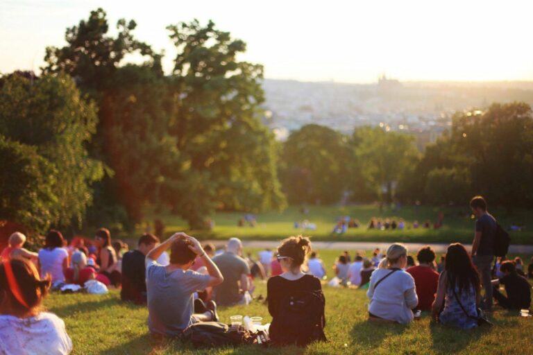 People picnicking on a grassy park hill at sunset, with a distant city skyline.