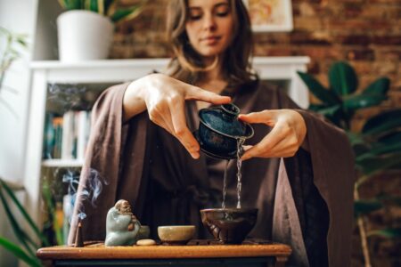 Woman in brown robes pours tea from a small dark teapot into a cup on a wooden table, with a smoking incense statue nearby and houseplants in the background.