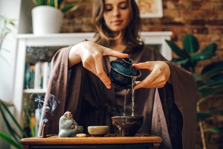 Woman in brown robes pours tea from a small dark teapot into a cup on a wooden table, with a smoking incense statue nearby and houseplants in the background.