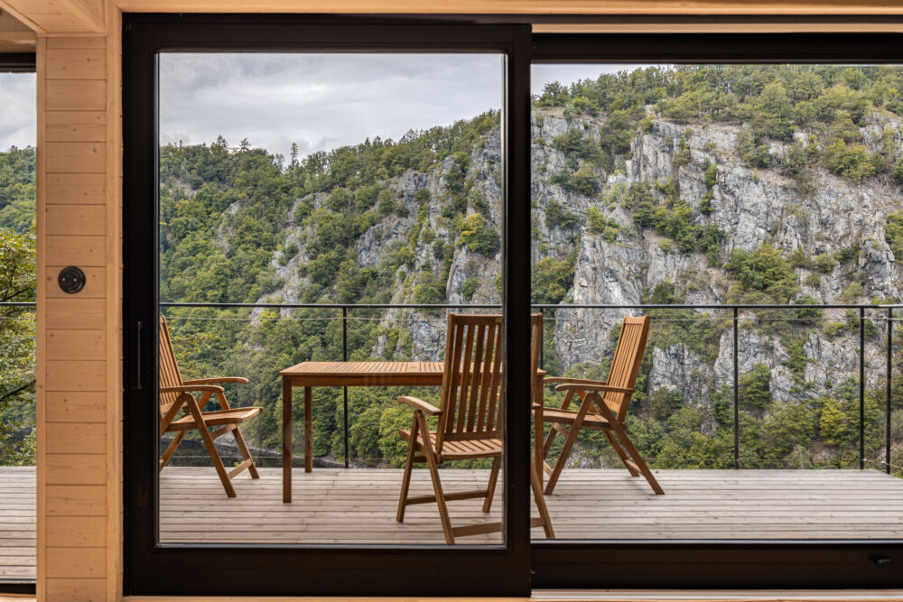 Wooden patio with chairs and a table overlooking a rocky, forested cliff seen through a large glass door frame