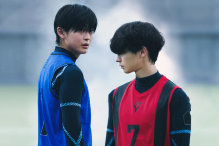 Two young athletes wearing blue and red jerseys stand face to face on a blurred field with a calm background.
