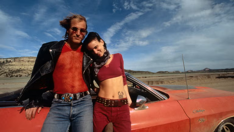Couple leaning on an orange vintage car in a desert landscape, dressed in 70s rock style with leather jacket and crop top.
