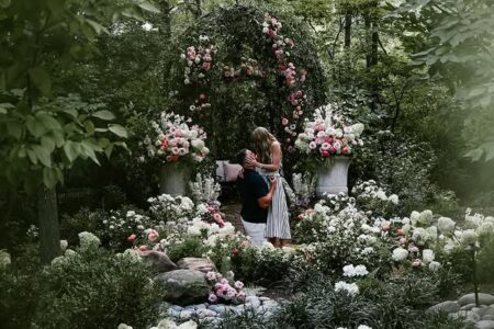 A couple stands in a lush garden under a rose arch, with pink and white flowers surrounding them. The man appears to be kneeling as the woman in a striped dress stands beside him, sharing a moment.