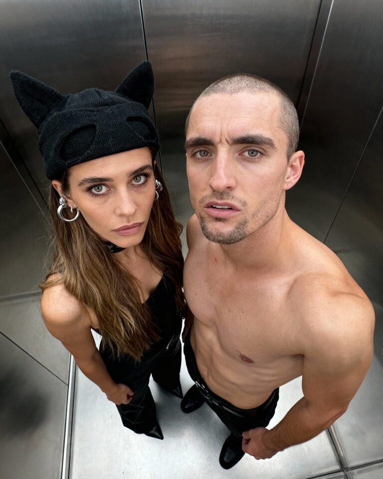 Two adults in an elevator posing for a selfie; woman wearing a black cat-ear hat and hoop earrings, man shirtless with stubble looking up at the camera.