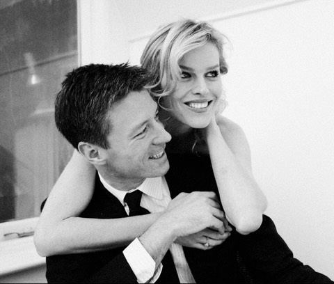 Black-and-white photo of a smiling couple, the woman wrapping her arms around the man's shoulders from behind.