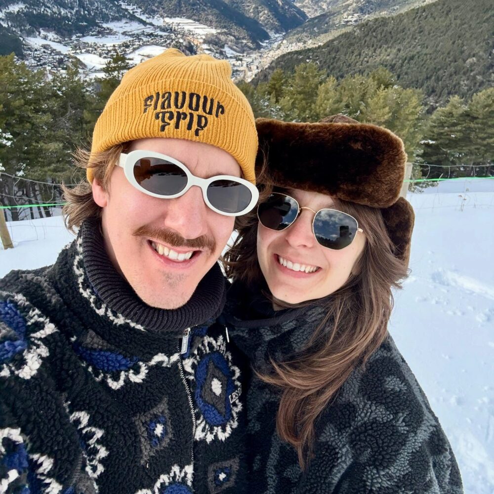 Smiling couple taking a selfie in a snowy mountain landscape, both wearing warm winter hats and sunglasses.
