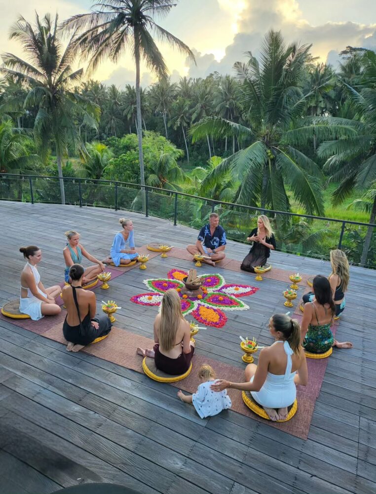 Group of people meditating on a wooden deck around a colorful flower mandala, with palm trees and tropical greenery in the background at sunset.