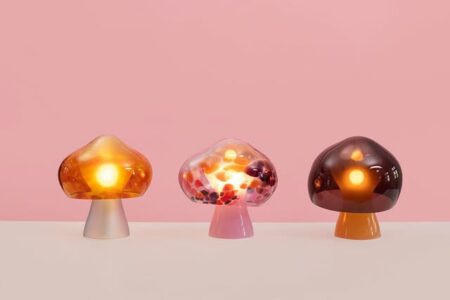 Three glass mushroom lamps glow amber, pink, and brown on a pale surface with a pink background.
