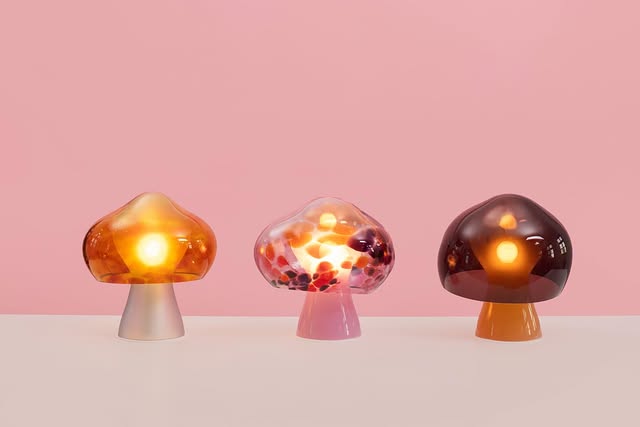 Three glass mushroom lamps glow amber, pink, and brown on a pale surface with a pink background.