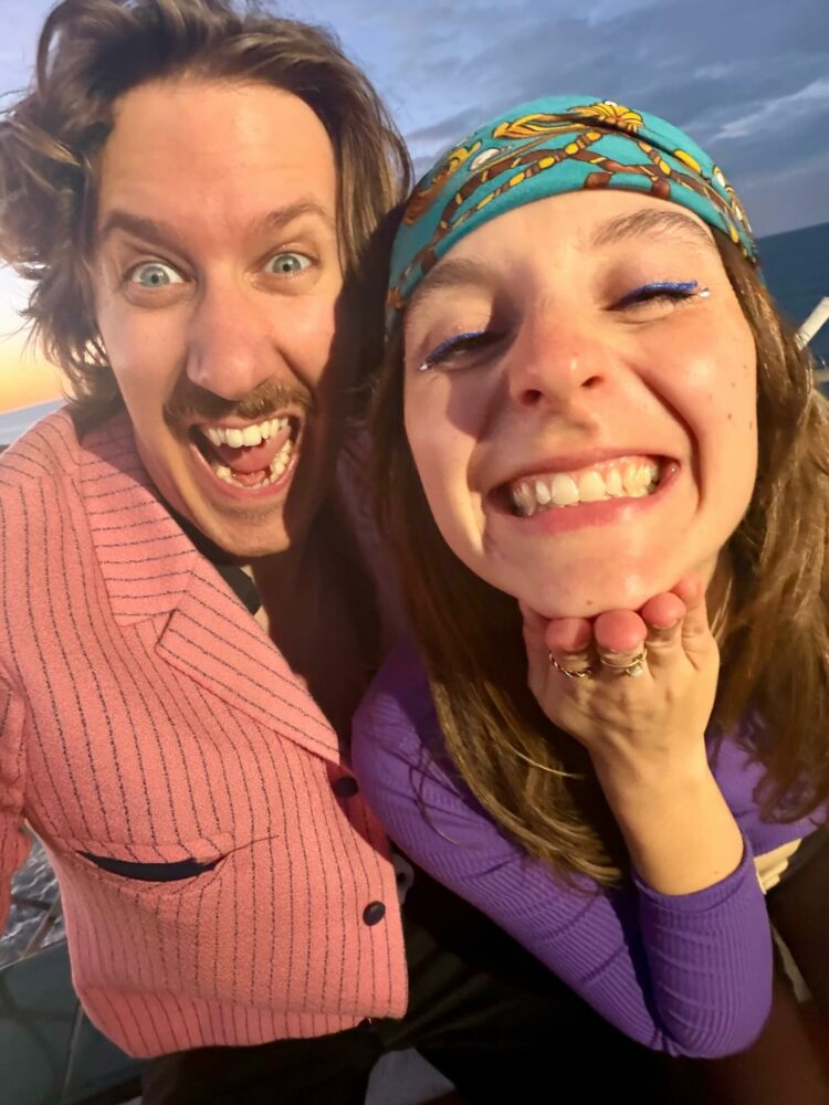 Close-up selfie of a man and woman at sunset, both smiling; the woman wears a teal patterned headband and blue eyeliner.