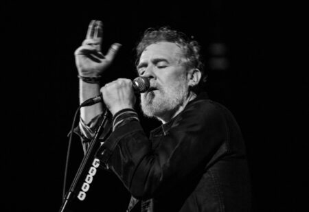 Black-and-white photo of a male singer on stage, eyes closed, singing into a microphone with his right hand, left hand raised.