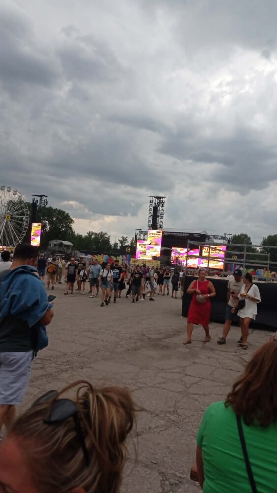 Outdoor music festival scene with a crowd around a main stage, large video screens, and a ferris wheel on the left under a cloudy sky