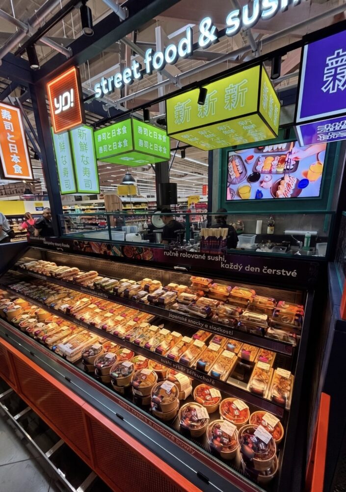 Sushi counter with refrigerated displays and assorted pre-packaged sushi trays, bright street food signs overhead in a market setting.