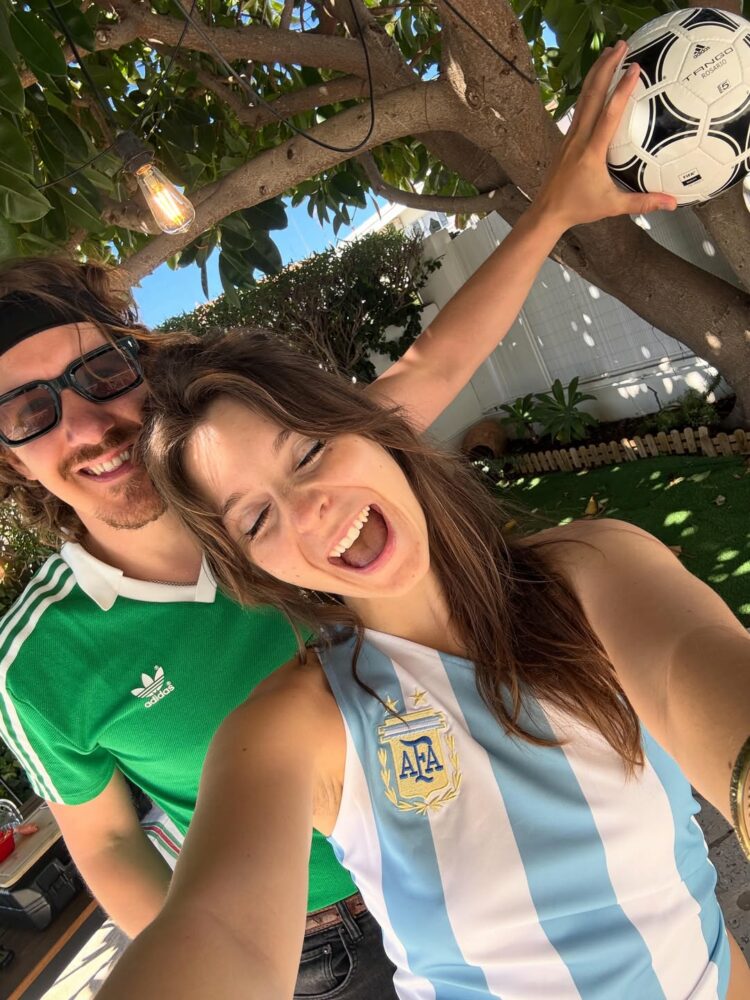 Two friends posing for a sunny outdoor selfie; a woman in an Argentina soccer jersey holds a soccer ball in a tree as a man in a green Adidas shirt smiles beside her.