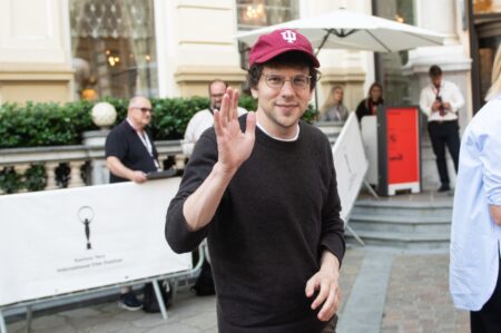 Man wearing a maroon cap with a white IU logo waves at the camera outdoors, with event barriers and onlookers in the background.