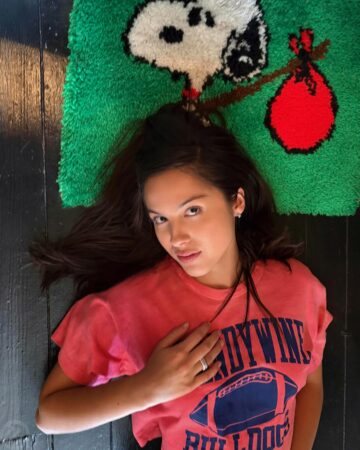 Woman lying on a dark wooden floor, head on a green Snoopy rug, wearing a pink T-shirt and a ring on her finger.