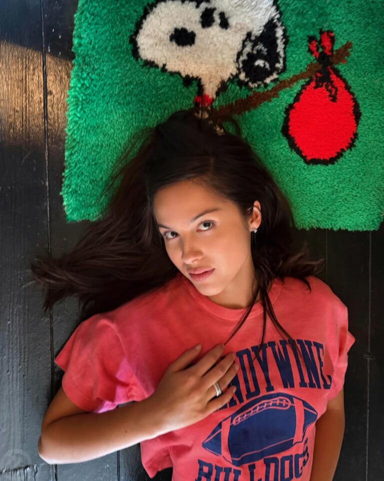 Woman lying on a dark wooden floor, head on a green Snoopy rug, wearing a pink T-shirt and a ring on her finger.