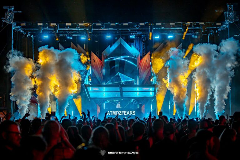 Crowd at a high-energy dance concert with blue geometric stage, fog, and orange flames rising from the sides.
