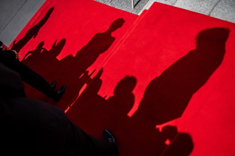Red carpet with dark silhouettes of guests standing and walking; photographer's shadow visible along the bottom-right edge.