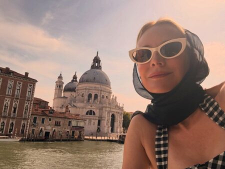 Woman in white cat-eye sunglasses and checkered scarf poses by a Venetian canal with the Santa Maria della Salute dome in the background, sunset sky behind her.