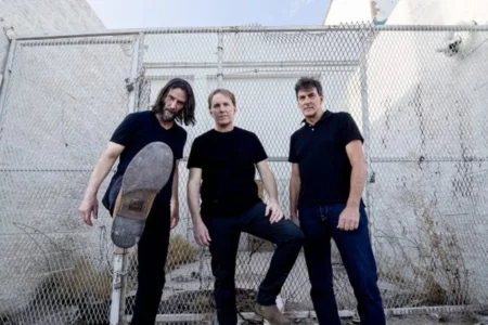 Three men in black shirts stand in front of a chain-link fence; the man on the left holds a skateboard resting on his leg.