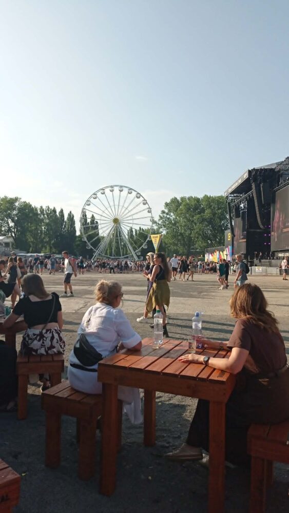 Outdoor festival scene with a large ferris wheel in the background, a stage on the right, and people seated at wooden picnic tables.